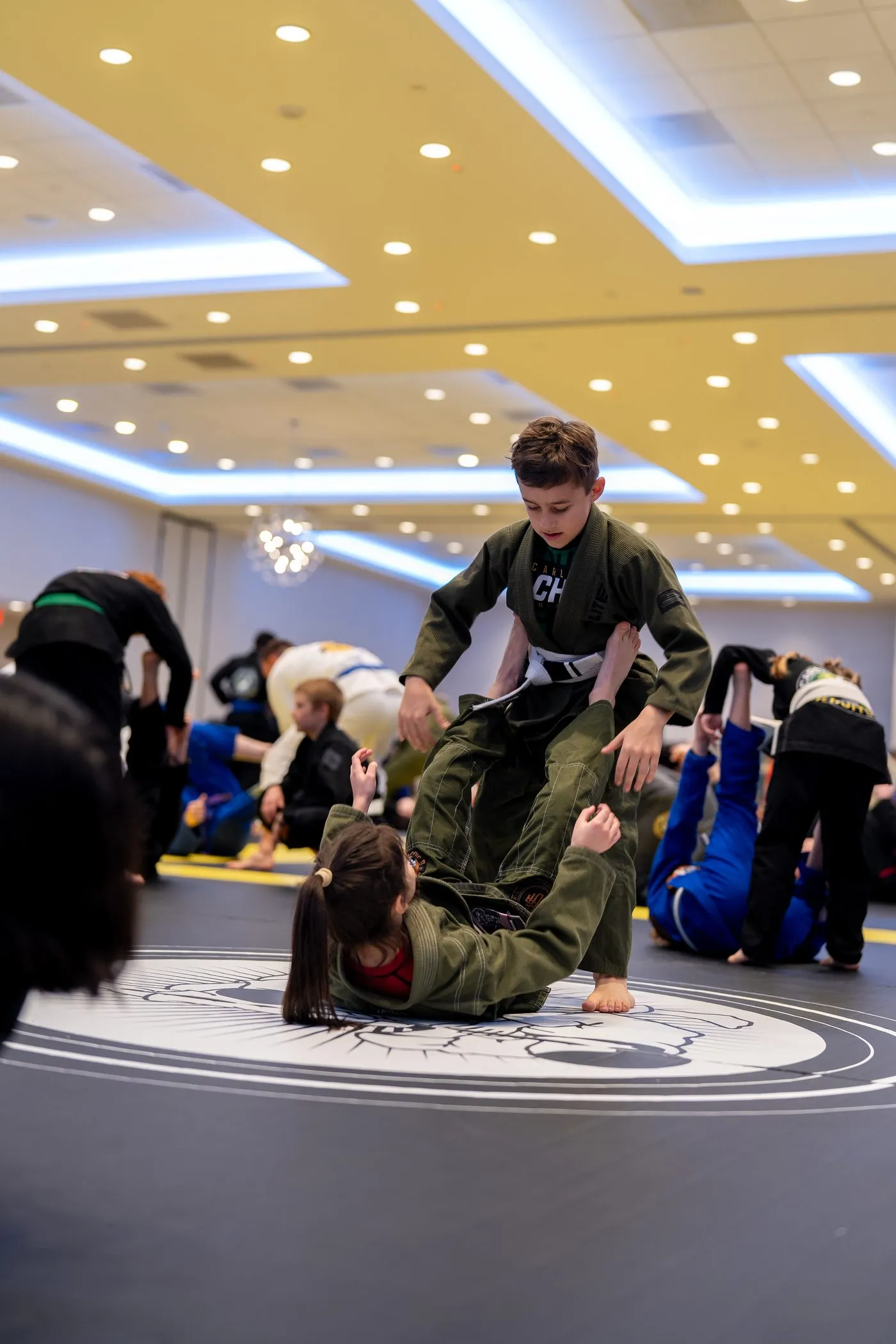 A young student in an olive gi working a technique on a partner on the mat