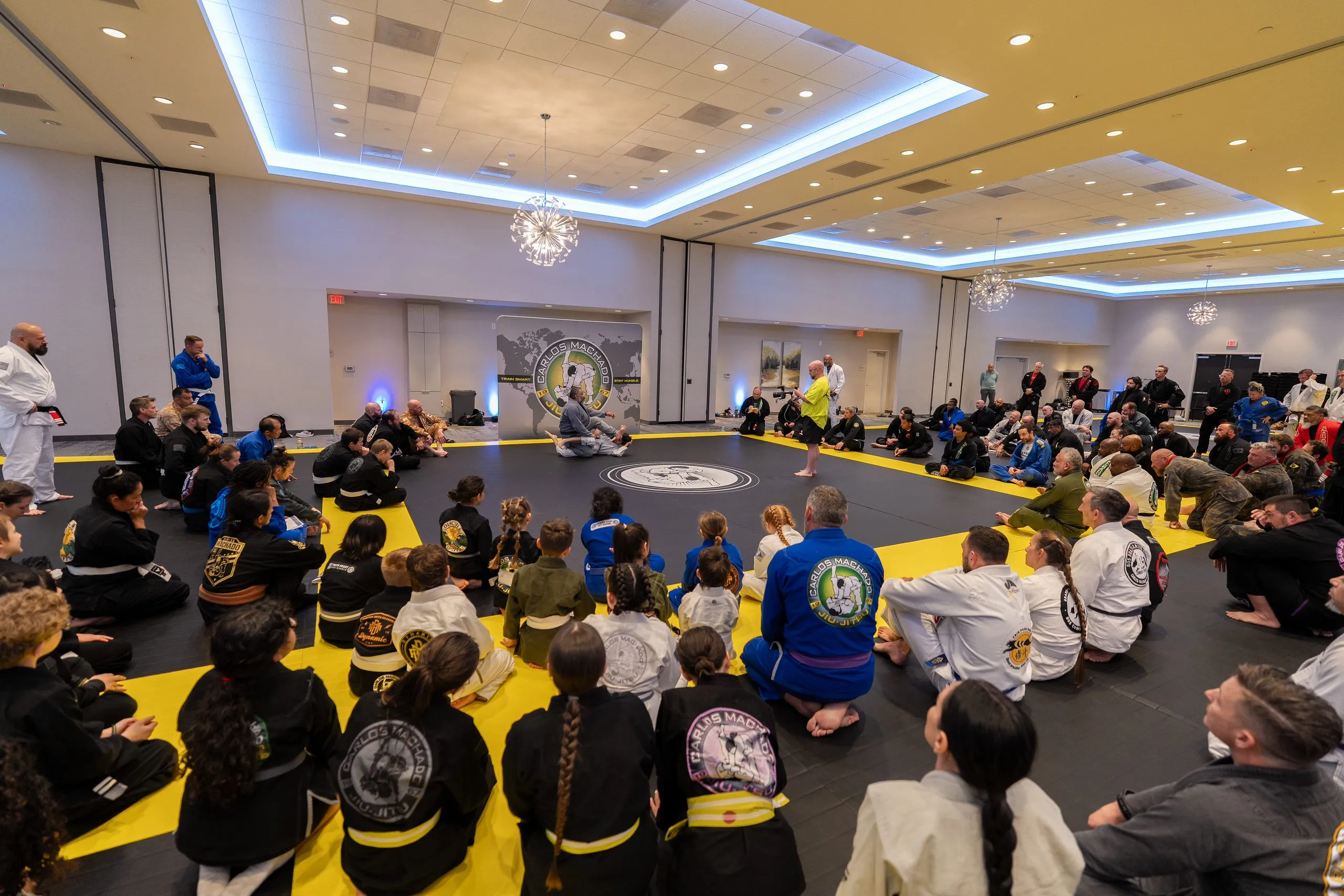 The full Summit seated in a circle around the Carlos Machado Jiu-Jitsu mat logo, adults and children together, the world-map banner behind them