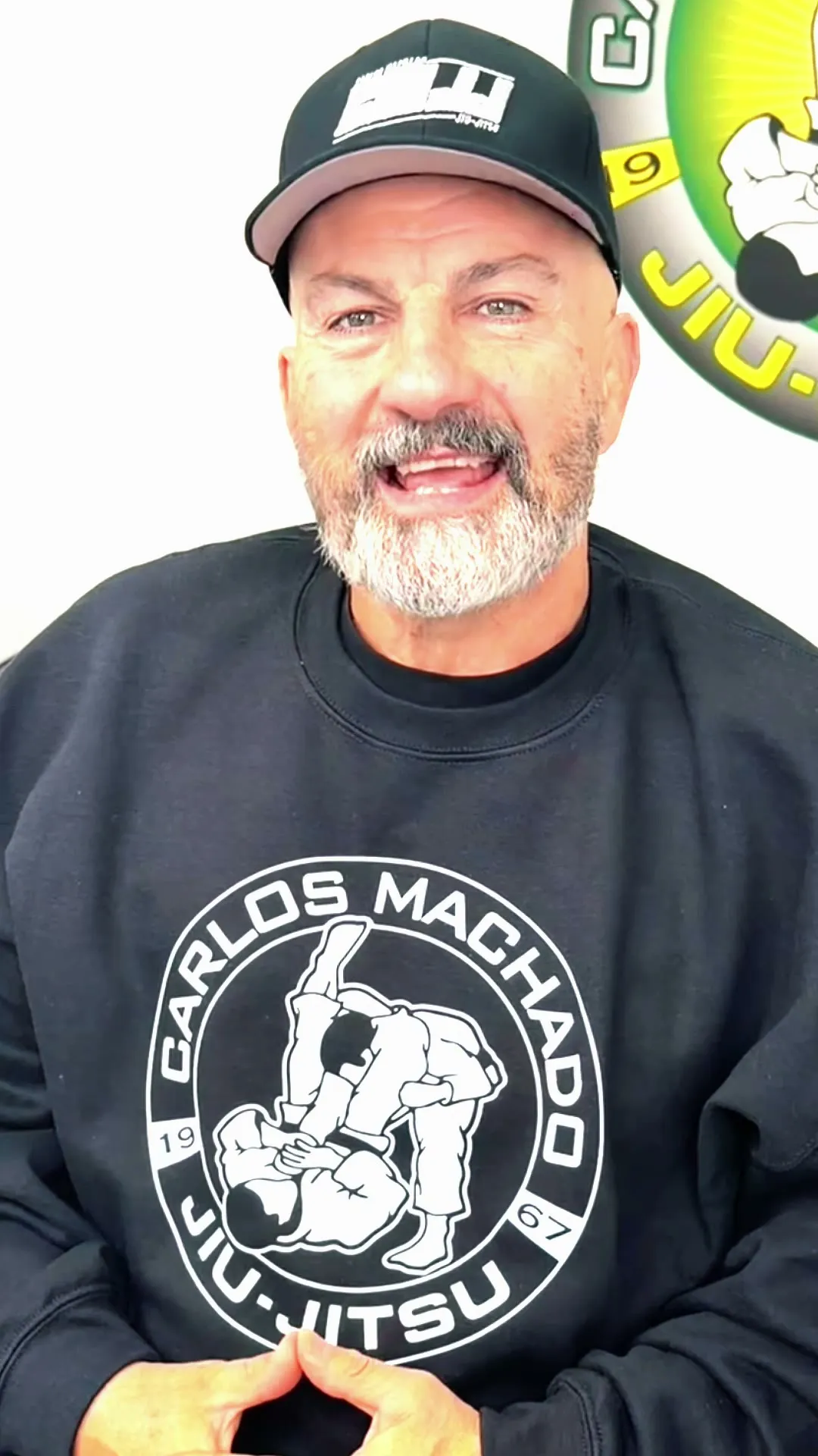 Master Carlos Machado speaking to camera in front of the Carlos Machado Jiu-Jitsu crest