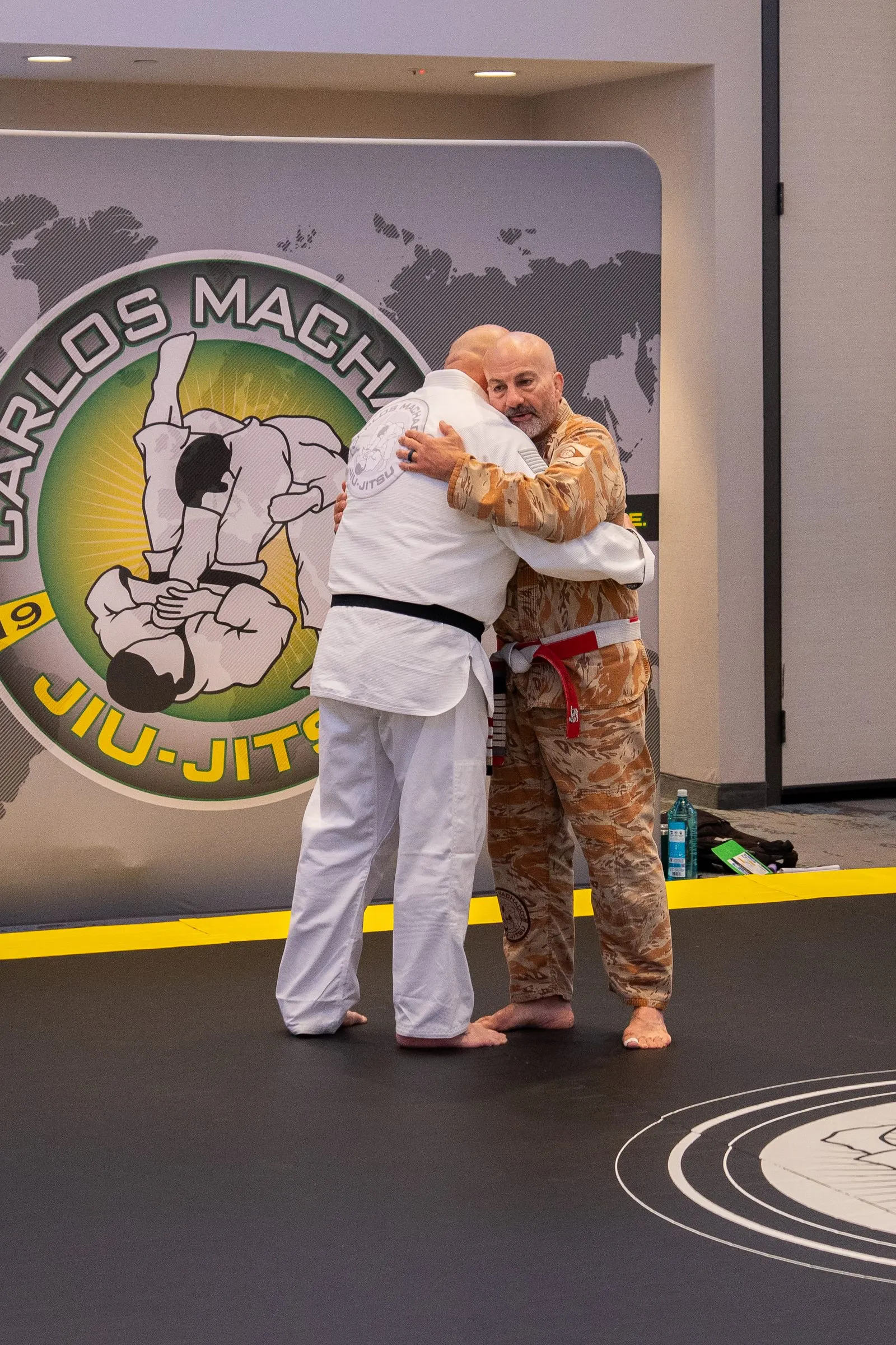 Two practitioners embracing on the mat immediately after a belt promotion, the Machado banner filling the frame behind them